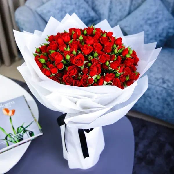 Crimson Deluxe 50 baby roses bouquet by Ward and Haneen wrapped in white paper, featuring vibrant red roses and delicate greenery.